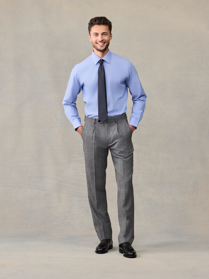 Man in a light blue shirt and navy tie with grey plaid trousers, hands in pockets.
