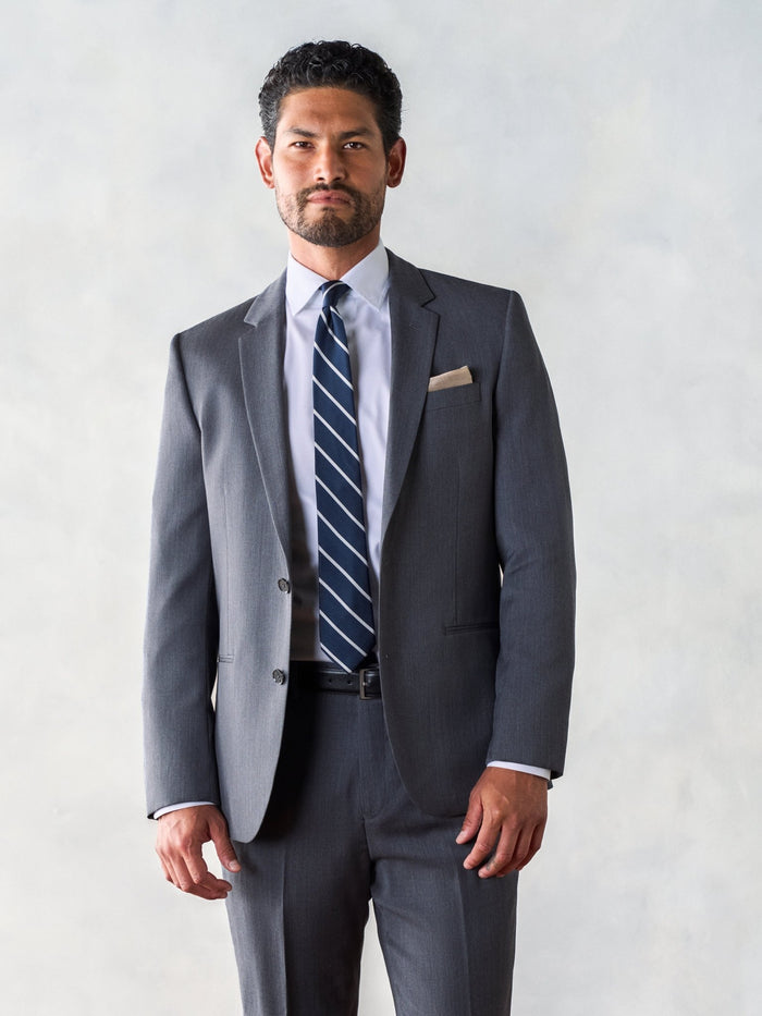 Man in a grey suit with a navy striped tie and pocket square, standing.