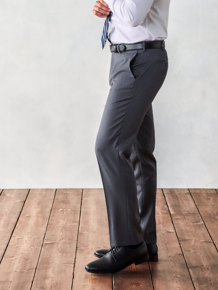 Side profile of grey suit trousers, black belt, and black shoes, hand adjusting navy tie