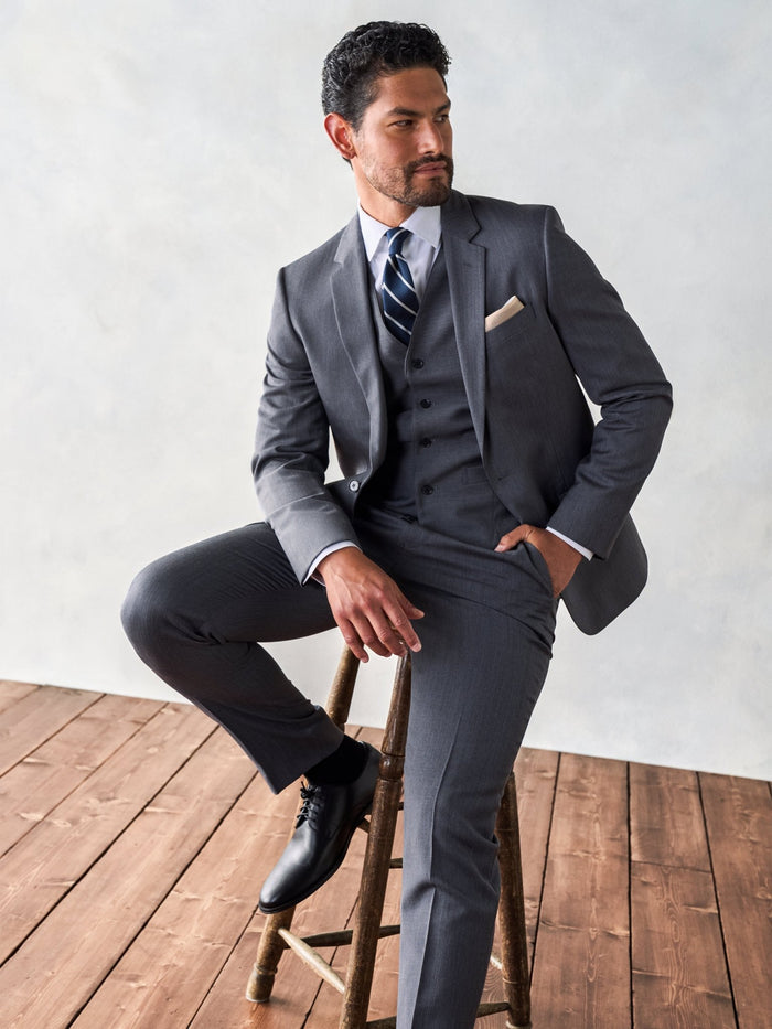 Man seated on wooden stool in three-piece grey suit, one leg propped, hand on knee