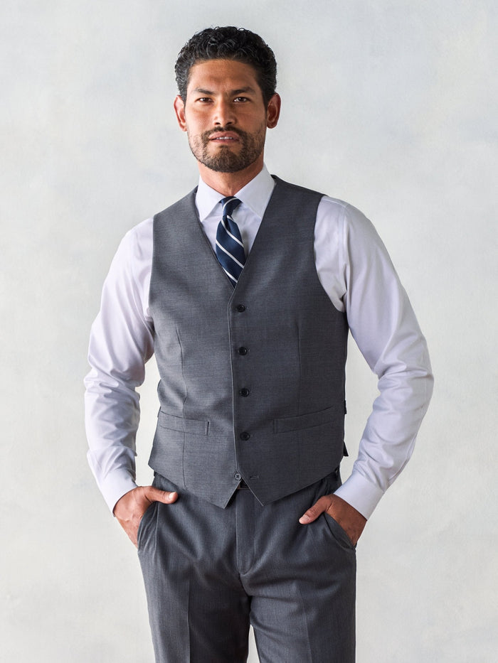 Man in grey vest and navy striped tie, hands in pockets, standing against grey backdrop