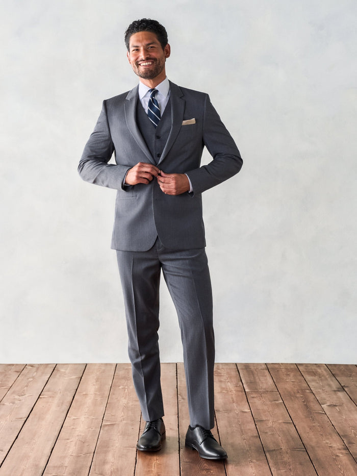 Smiling man buttoning grey suit jacket over vest, standing on wood floor