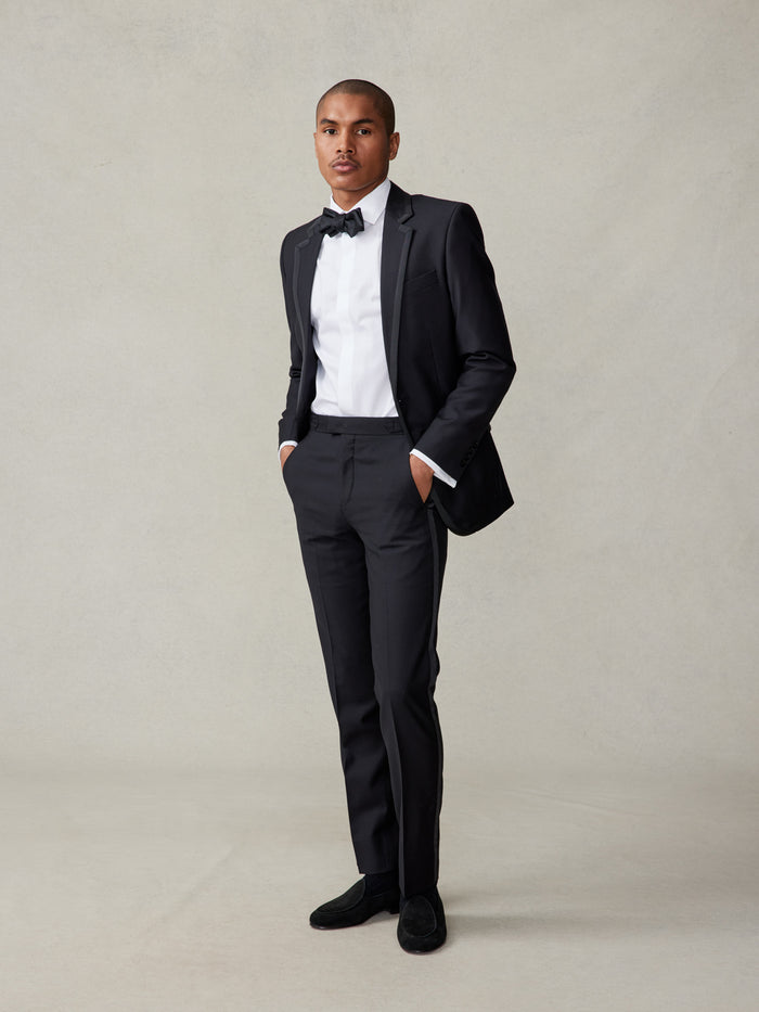 Man in black tuxedo and bow tie, one hand in pocket, standing full-length