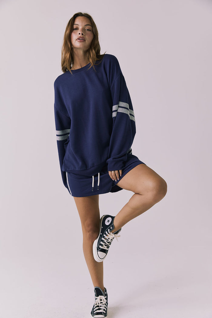 Full body front view of a woman wearing Chaser's navy blue Cornell Sapphire & Heather Grey Pullover with grey stripes on sleeves and matching Hanoverr shorts on a plain background