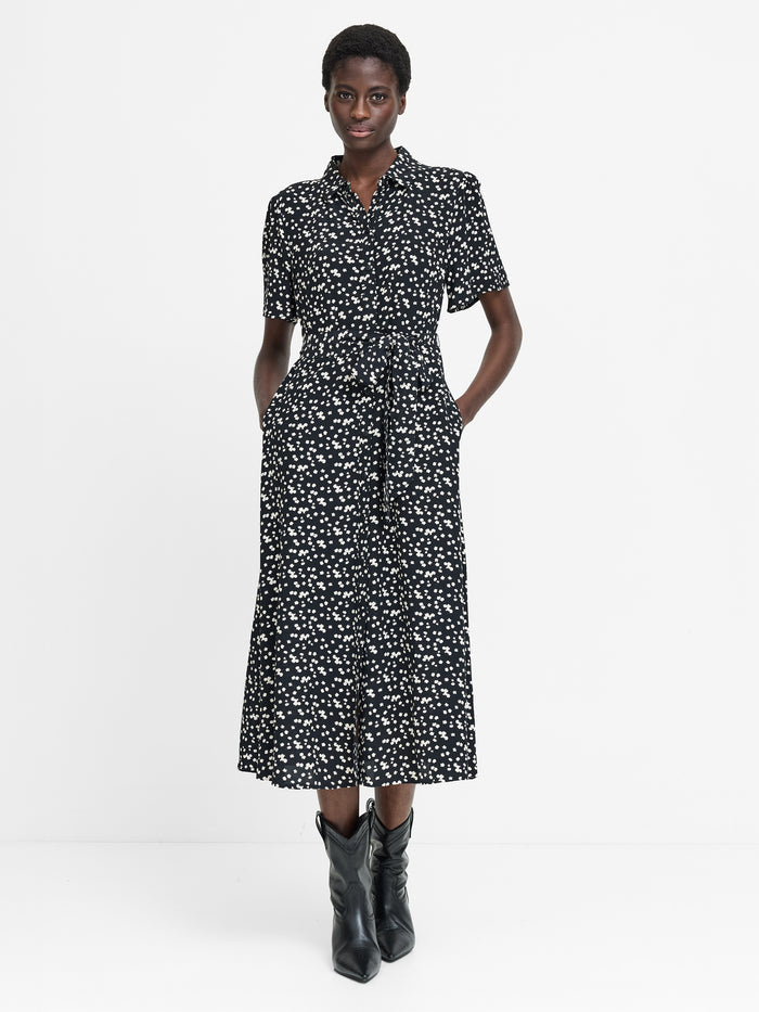 A woman wearing a short sleeved black midi length dress with a shirt collar, button down fastening, self-tie drawstring waist, two pockets and a white floral pattern.