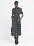 A woman wearing a short sleeved black midi length dress with a shirt collar, button down fastening, self-tie drawstring waist, two pockets and a white floral pattern.