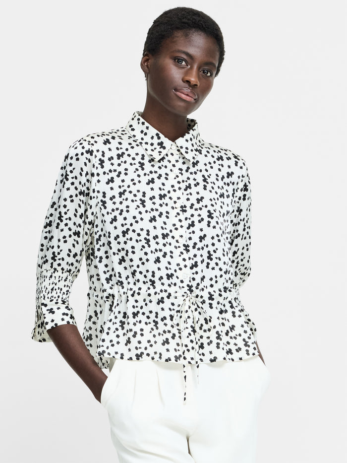 A woman wearing a cream button down shirt with a black floral pattern, 3/4 length sleeves, a collared neckline, and a self tie drawstring waist.