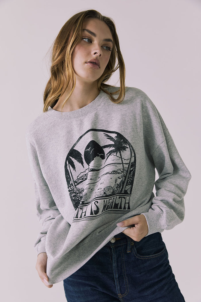 Front view of a woman pulling the hem of Chaser's gray Health Is Wealth Long Sleeve with a graphic design on a plain background