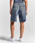 A person shown from the back wearing medium-wash blue denim shorts with a raw frayed hem, paired with white sneakers against a light gray studio background.