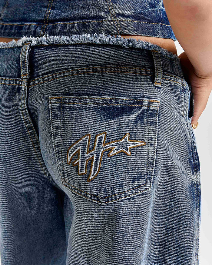 Close-up back view of a person wearing blue denim jeans with a frayed waistband and a large embroidered monogram logo on the back pocket.
