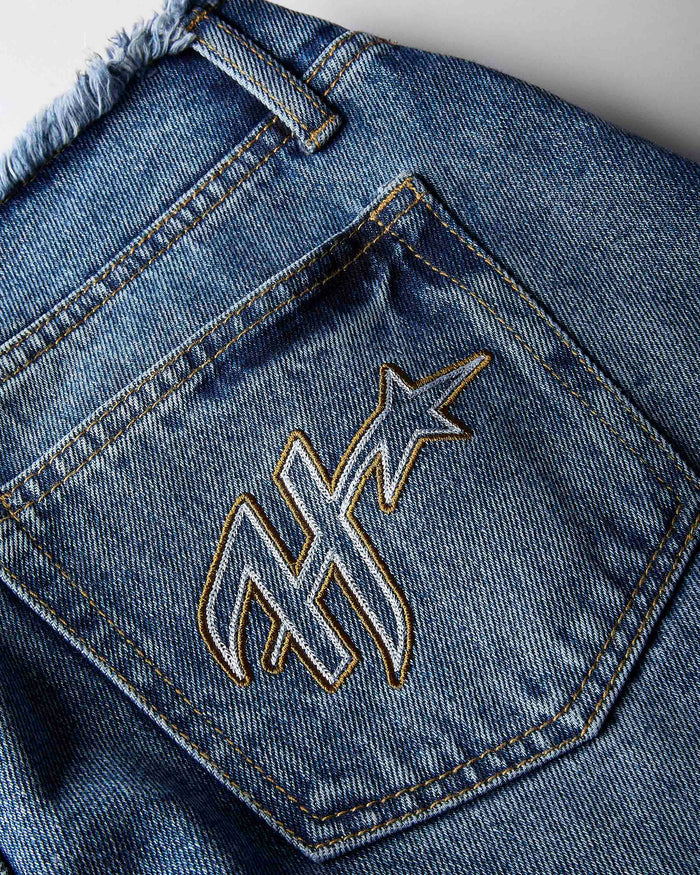 Close-up of the back pocket of blue denim jeans with a large embroidered monogram logo outlined in gold thread.