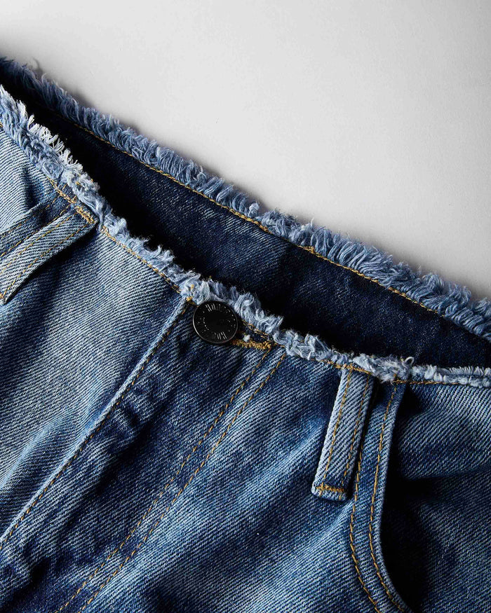 Close-up of a pair of blue denim jeans showing the raw, frayed waistband, a single button closure, and belt loops against a light gray background.