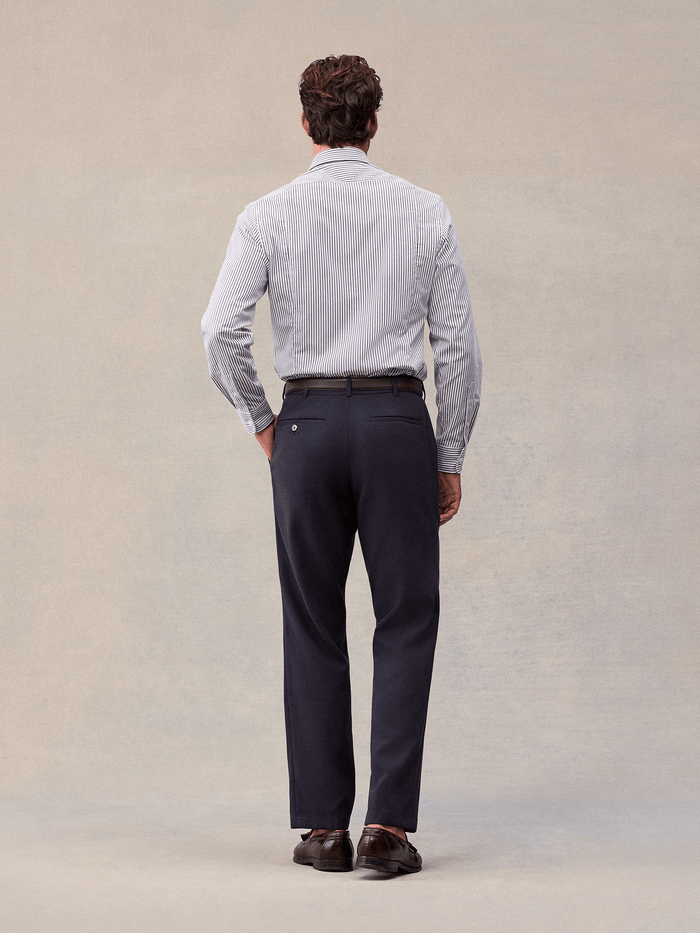 A man in admiral navy suit pants and a striped shirt, back view, full length, beige backdrop.