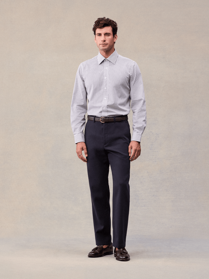 A man in admiral navy suit pants and a striped shirt, full length front view, beige backdrop.