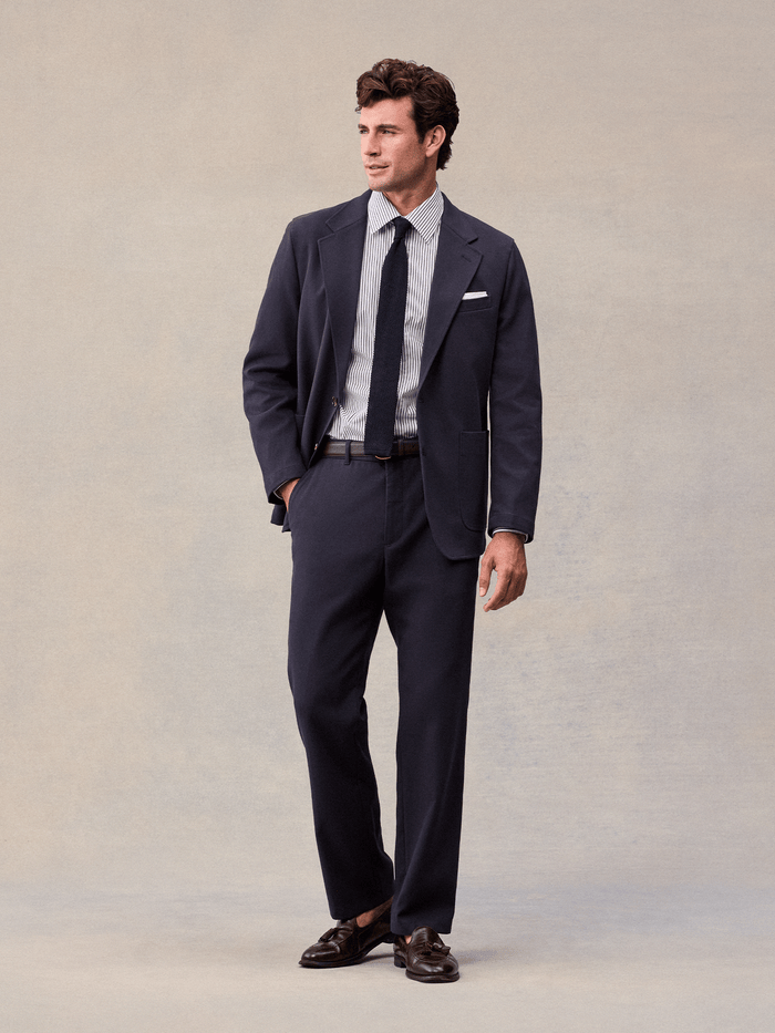 A man in an admiral navy suit with a navy knit tie and brown tassel loafers, full length front.