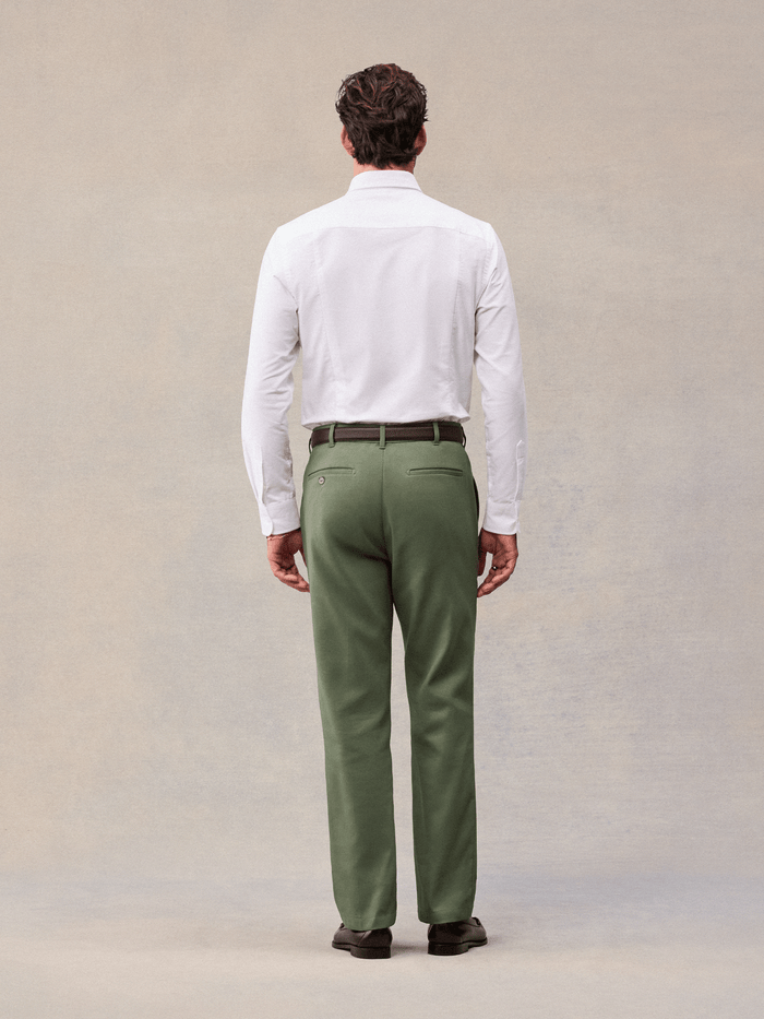 A man in dusty sage suit pants and a white shirt, back view, full length, beige backdrop.