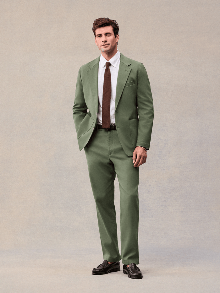 A man in a dusty sage suit with a brown knit tie and brown penny loafers, full length front.