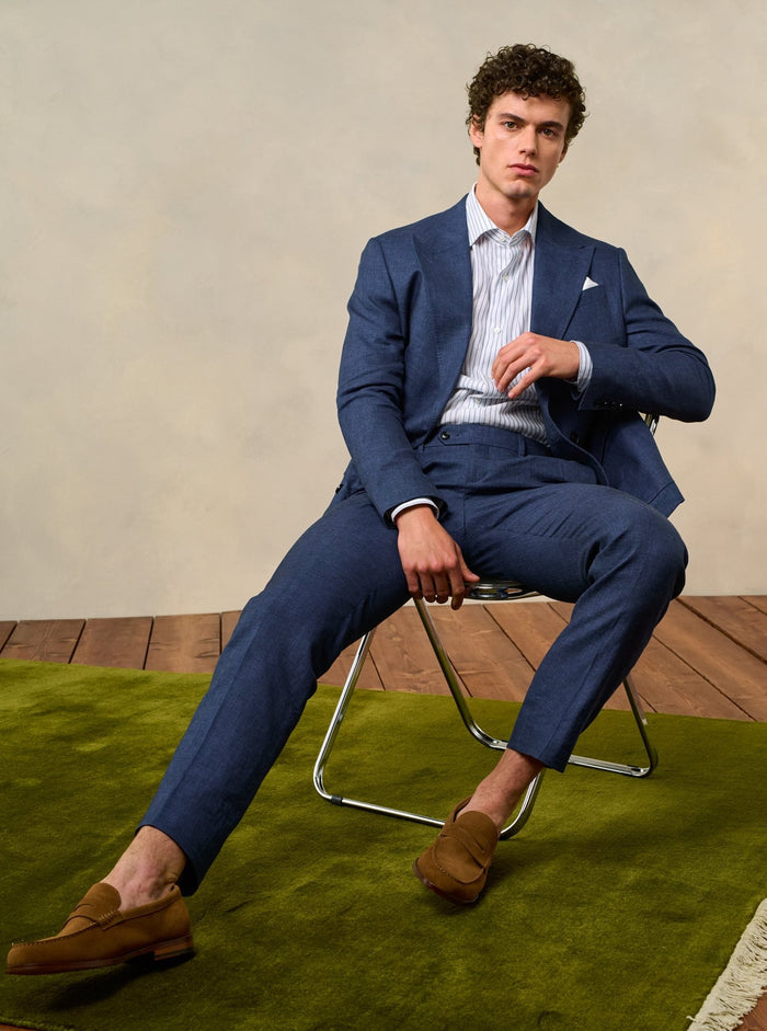 Man seated on a wire chair in an ink blue linen suit with tan suede loafers
