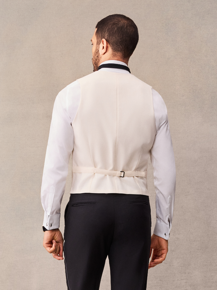 Back view of man in ivory tuxedo vest with adjustable strap buckle over black trousers