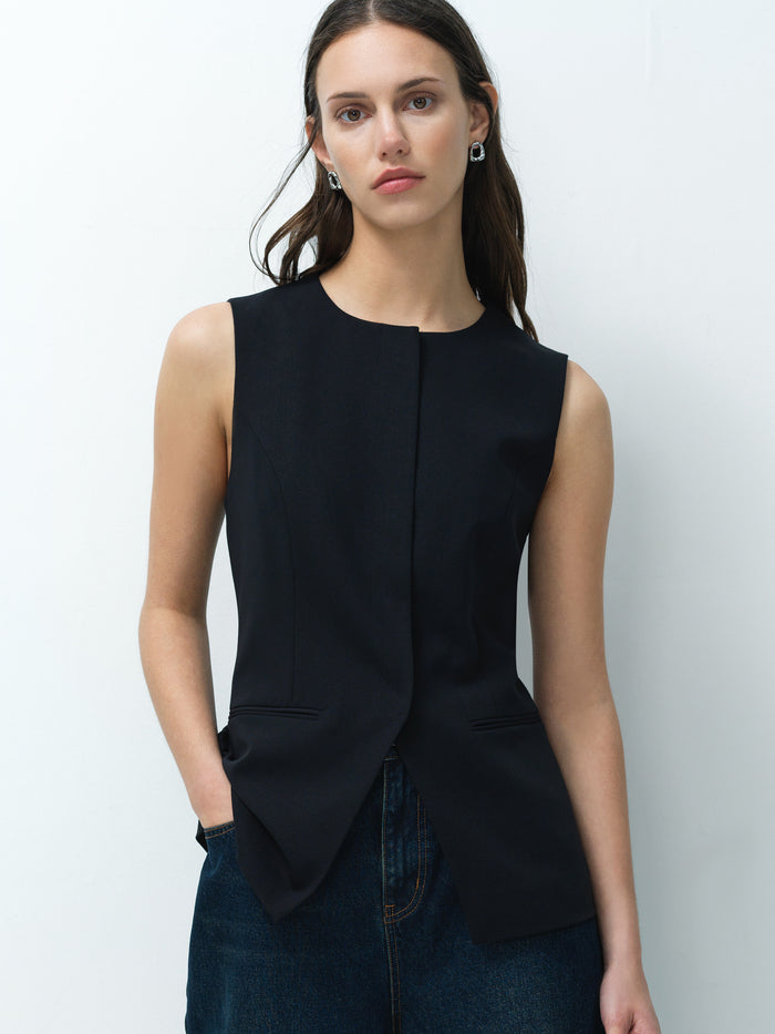 A woman wearing a black tailored long line waistcoat with front pockets, a round neckline and concealed popper fastening.