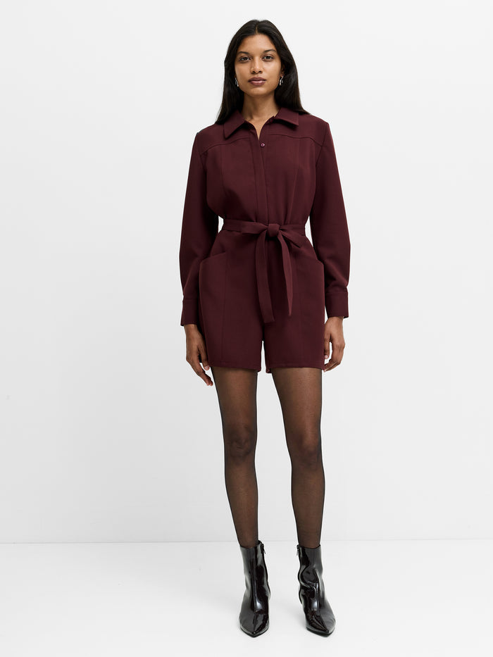 A woman is wearing a long sleeve burgundy playsuit featuring a collared neckline, a button down fastening and a self tie belt.