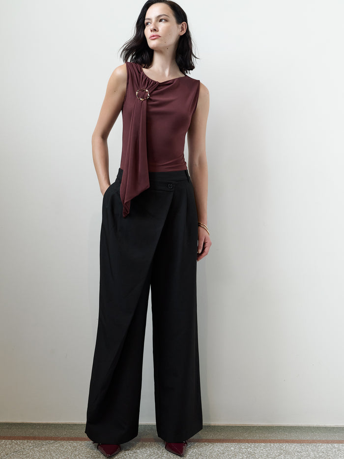 A woman wearing black high waisted Trousers with an a asymmetric waistband front overlay, front pleats, 2 side pockets, a wide leg Full length tailored full length silhouette.
