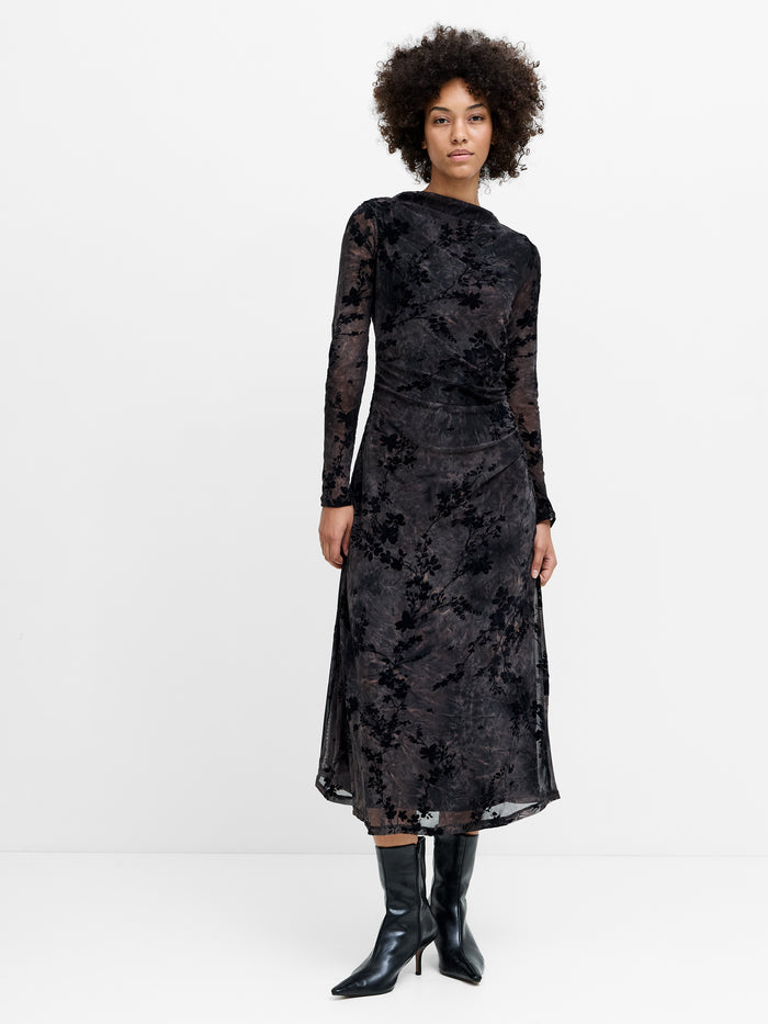 A woman wearing a long sleeved mesh midi dress featuring a black and brown floral print, a boat neckline, and gathered side detailing.