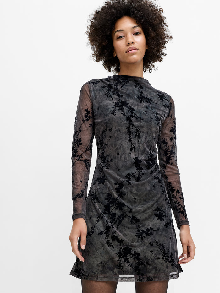 A woman wearing a mesh floral brown and black mini dress with semi sheer sleeves and gathered side detailing.