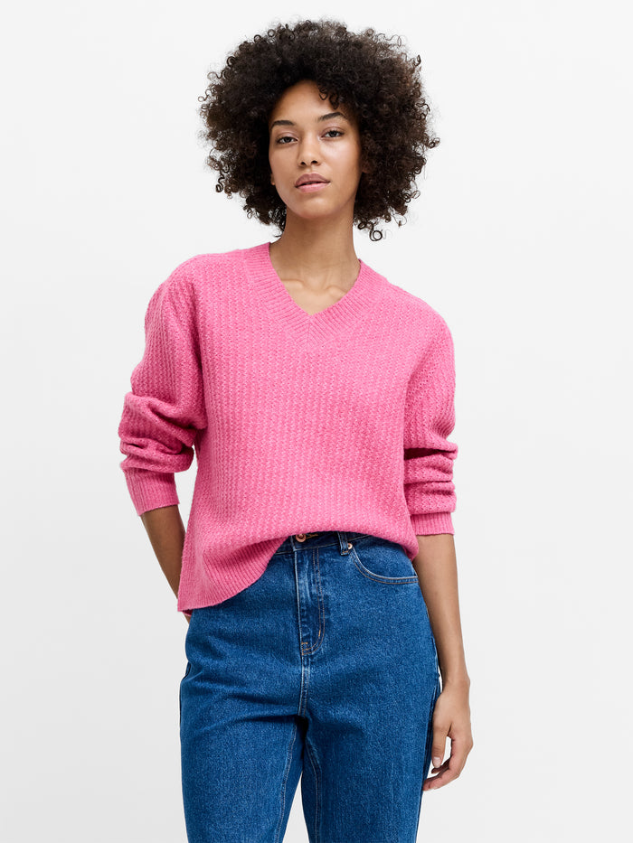 A woman wearing a pink textured knit jumper with a v-neckline, long sleeves and a relaxed fit.