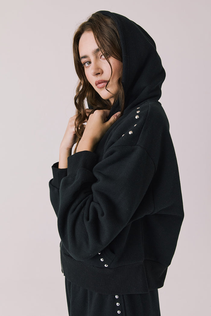Side view of woman wearing Chaser’s Kiki True Black Zip Up Hoodie with sequin embellishments, hood up, and matching Daze True Black Joggers on a white background