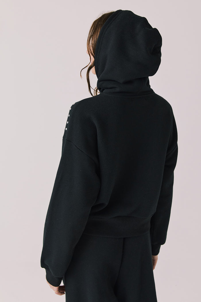 Back view of woman wearing Chaser’s Kiki True Black Zip Up Hoodie with sequin accents with hood up on the shoulders and pockets on a white background