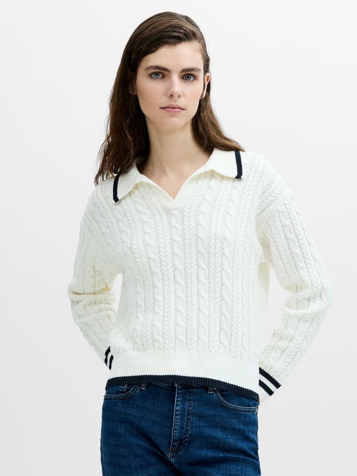 A woman wearing a white cable knit polo jumper with a V-neck, long sleeves and a navy contrast trim.