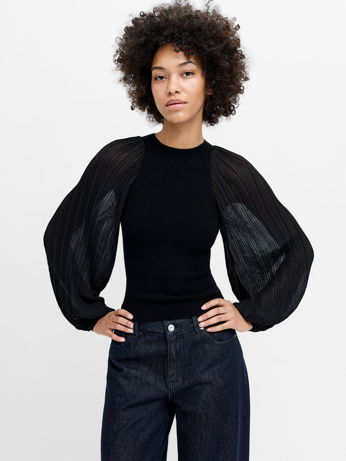 A woman wearing a black ribbed knit top with a crew neckline, voluminous pleated sleeves, and a fitted silhouette.