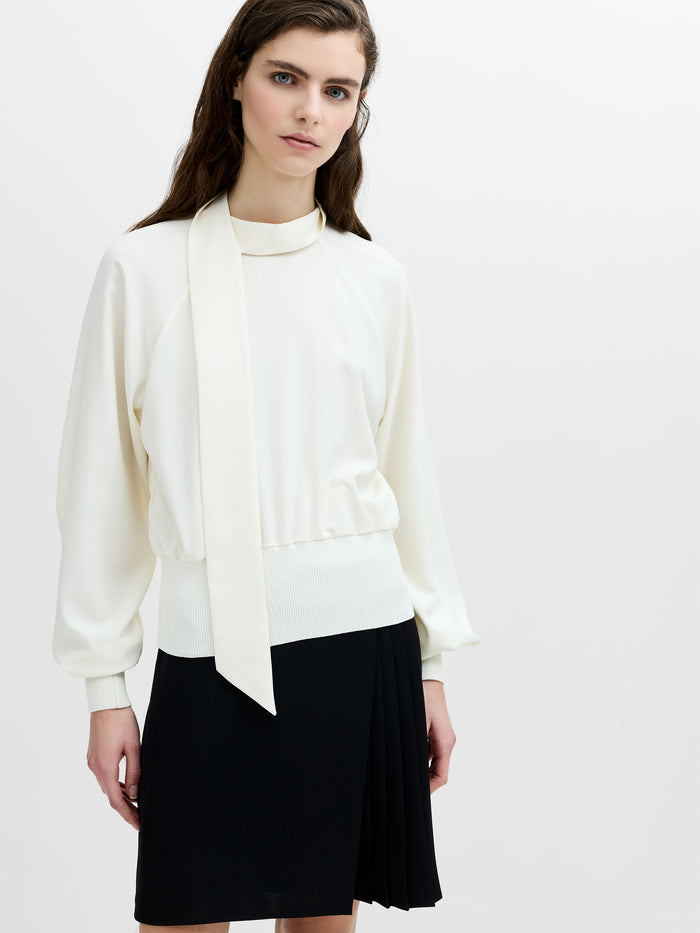 A woman wearing a cream long sleeved top with a draped scarf neckline, ribbed cuffs and a ribbed hem.