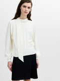 A woman wearing a cream long sleeved top with a draped scarf neckline, ribbed cuffs and a ribbed hem.