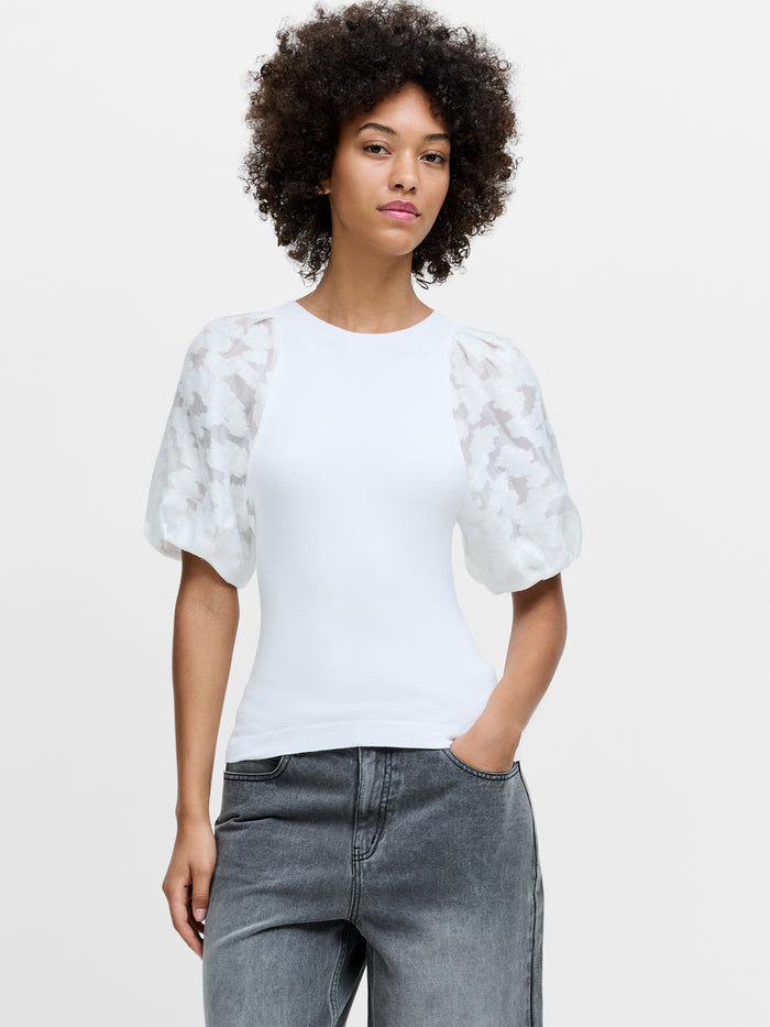 A woman wearing a white knit top with sheer puff sleeves, a crew neckline and a ribbed hem.
