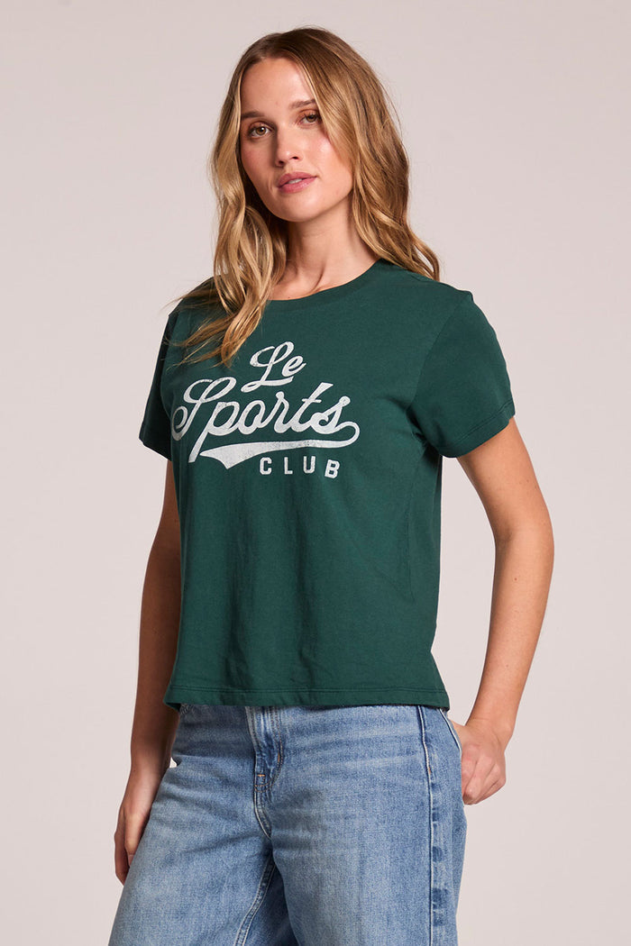 Side angle view of a woman wearing the Chaser Le Sports Club Tee on a neutral background with one hand on her hip