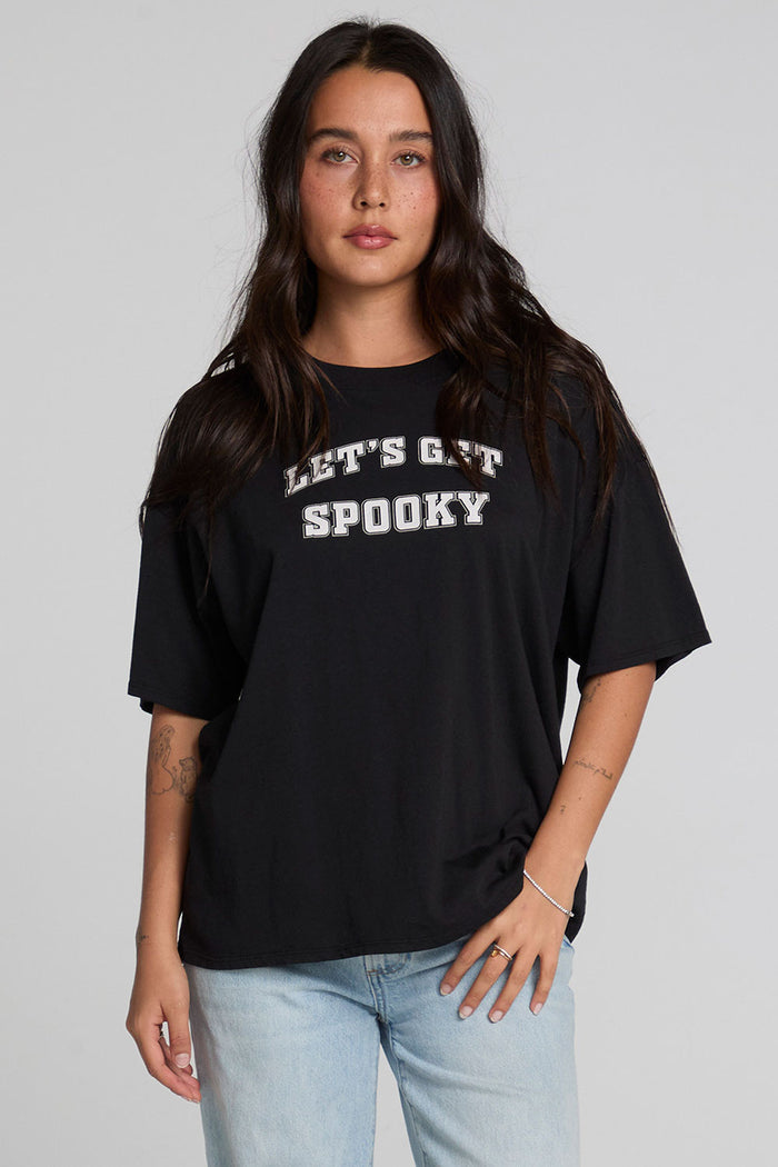 Front view of a woman wearing the Chaser Let's Get Spooky Tee on a neutral background with jeans and hands on leg