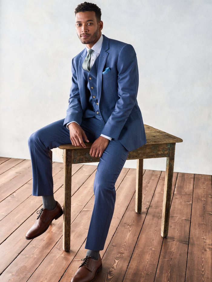 Man in light blue three-piece suit, sage tie, seated on wooden bench, brown shoes