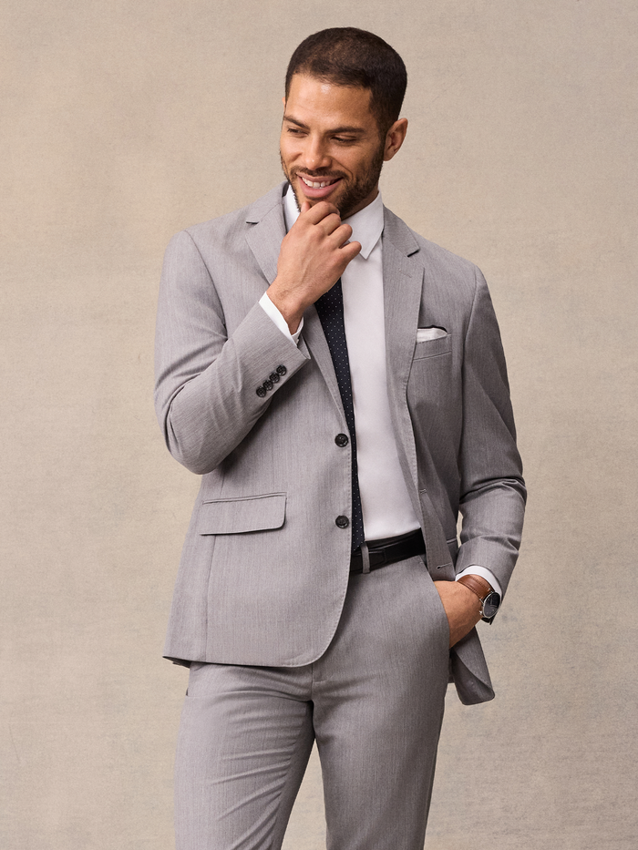 Man in light grey suit, navy pindot tie, hand at chin, smiling