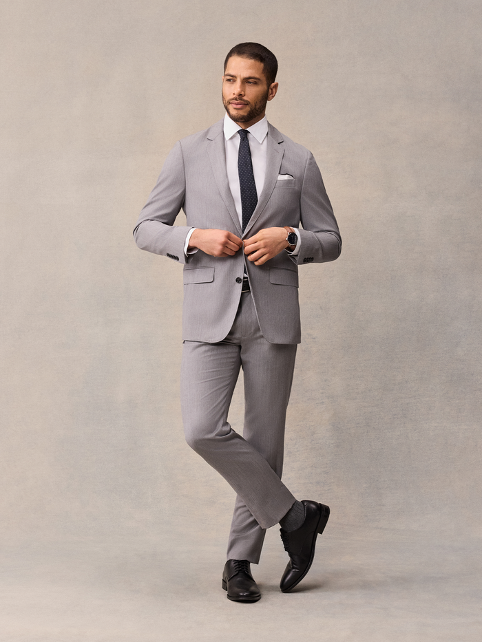 Man in light grey suit jacket, black pindot tie, buttoning jacket, walking pose