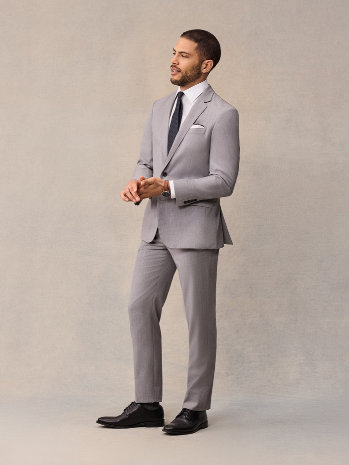 Man in light grey suit, navy pindot tie, three-quarter turn, hands clasped