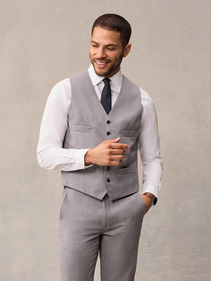 Smiling man in light grey suit vest, black pindot tie, hand raised