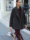 A woman wearing a dark grey coat with drop shoulders, long sleeves, an over sized lapel collar and a bomber fit.