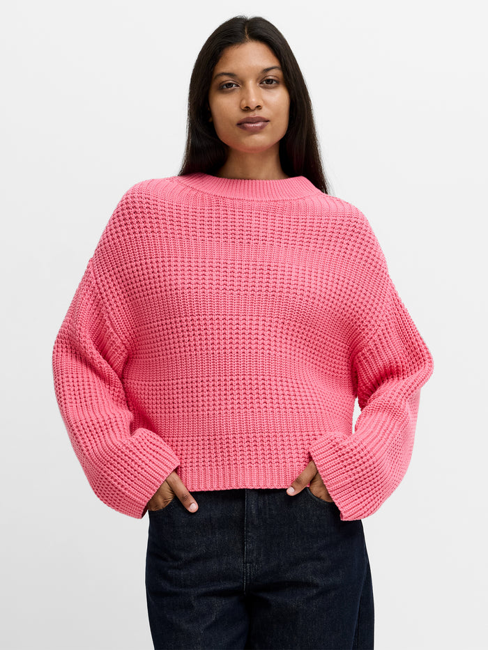 A woman wearing a textured knit Jumper with cropped shoulders, long wide sleeves, a mock neckline and a boxy fit.
