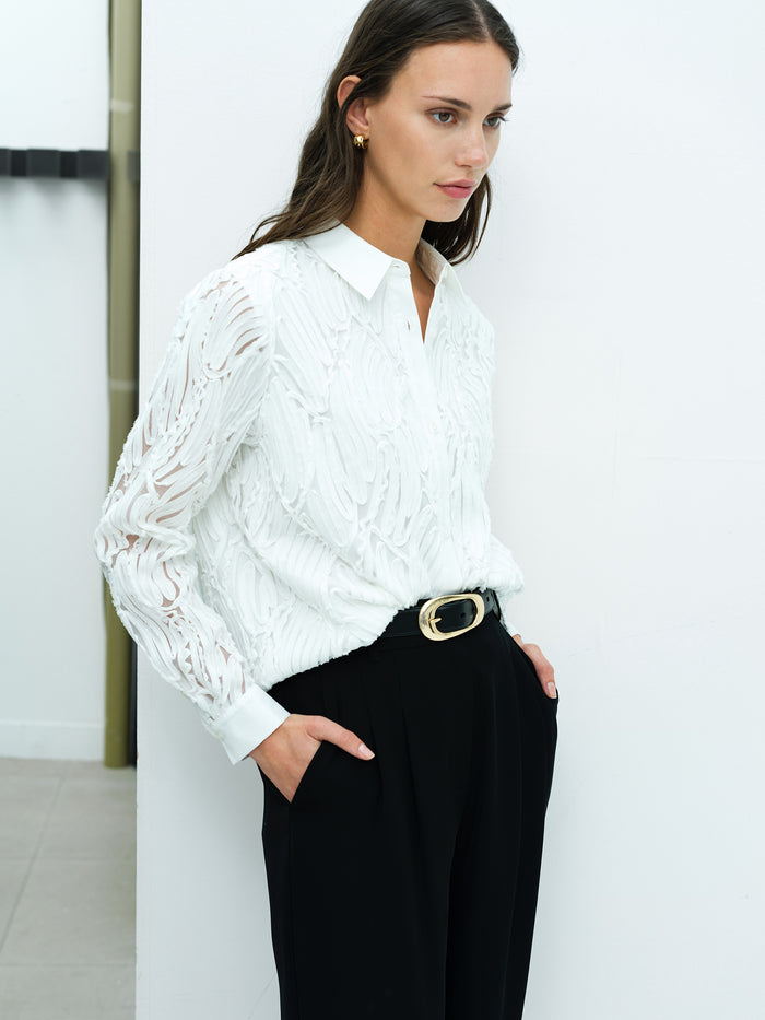 A woman wearing a white 3D textured long sleeve shirt with a pointed collar, button through fastening and semi-sheer sleeves.