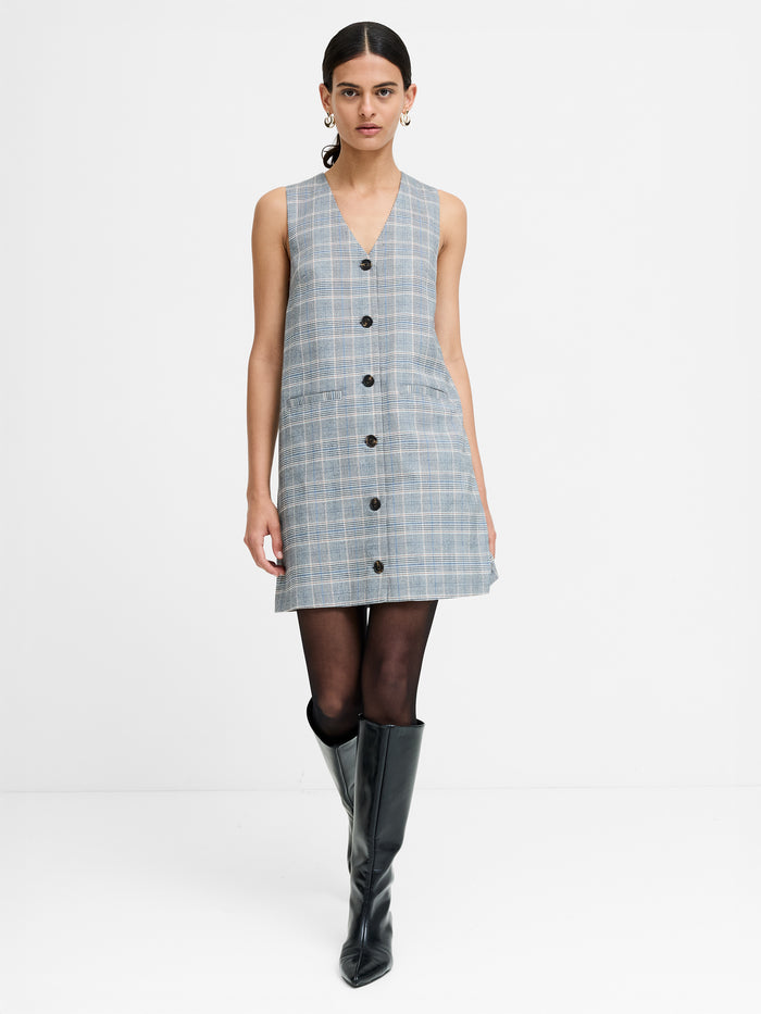 A woman wearing a sleeveless grey plaid dress with a button-through fastening, two pockets and a V-neck.