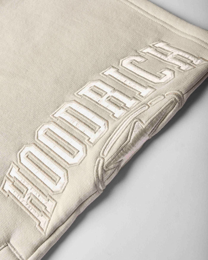 Close-up of a light beige or cream-colored fabric item, likely a sweatshirt or jacket, featuring large raised collegiate-style embroidery reading 'HOODRICH' across the surface.