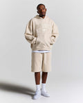 A man modeling an oversized beige hoodie with 'HOODRICH' text and a matching beige drawstring shorts set, styled with white t-shirt layers, white socks, and white sneakers against a light gray studio background.