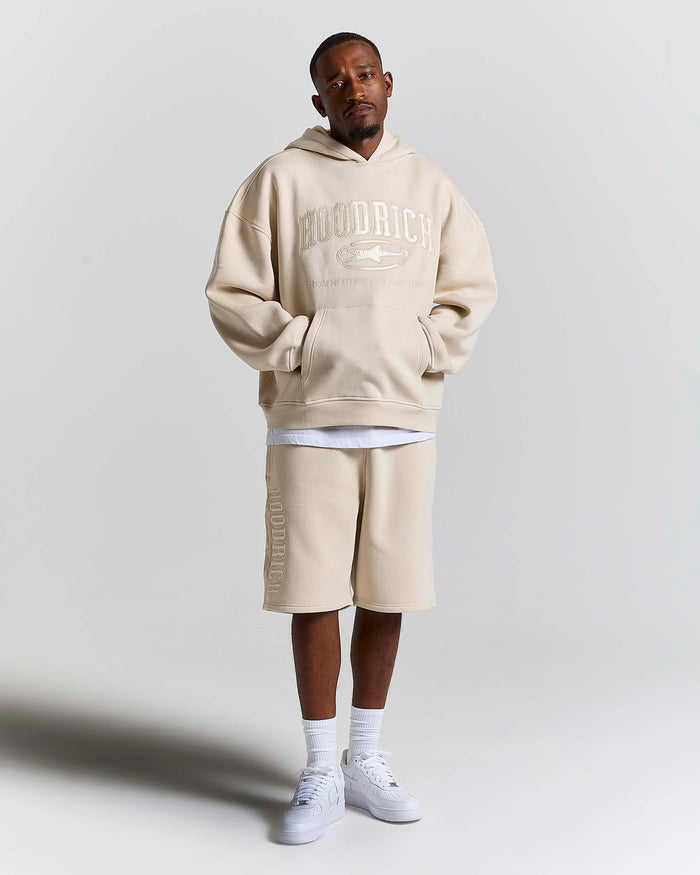 A man modeling an oversized beige hoodie with 'HOODRICH' text and a matching beige drawstring shorts set, styled with white t-shirt layers, white socks, and white sneakers against a light gray studio background.